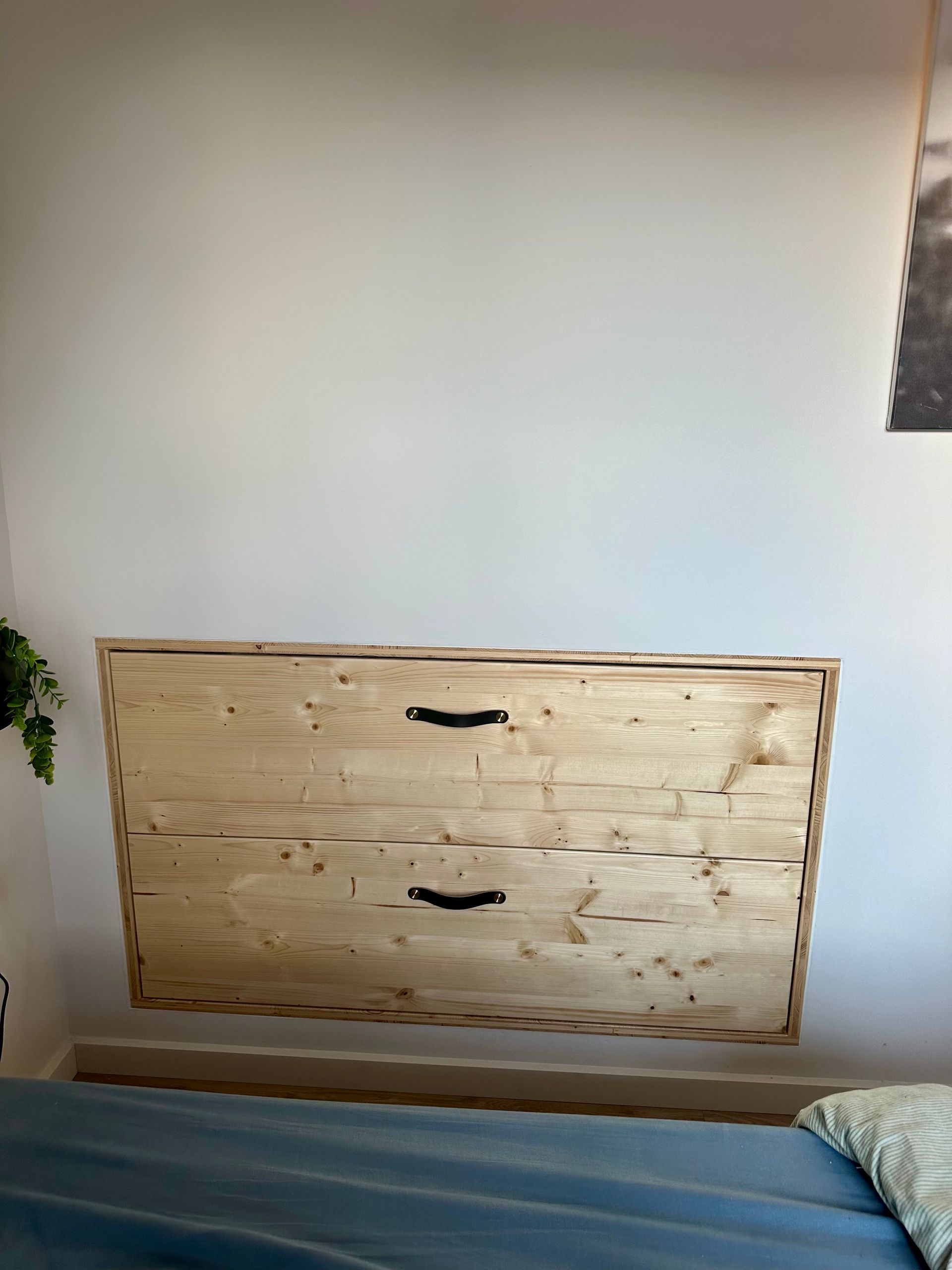 Commode en bois encastrée dans une chambre, sous un mur blanc et au-dessus d'un couvre-lit bleu.