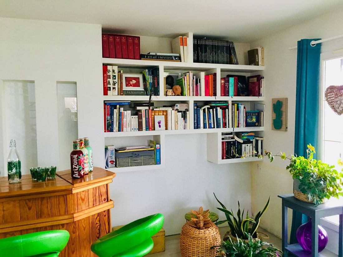 Salon lumineux avec bibliothèque murale, armoire en bois, plantes et chaises vertes près d'un rideau bleu.