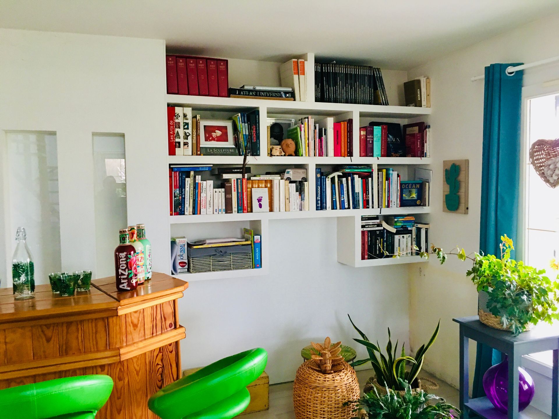 Salon lumineux avec étagères murales, armoire en bois, chaises vertes et plantes près d'un rideau bleu.