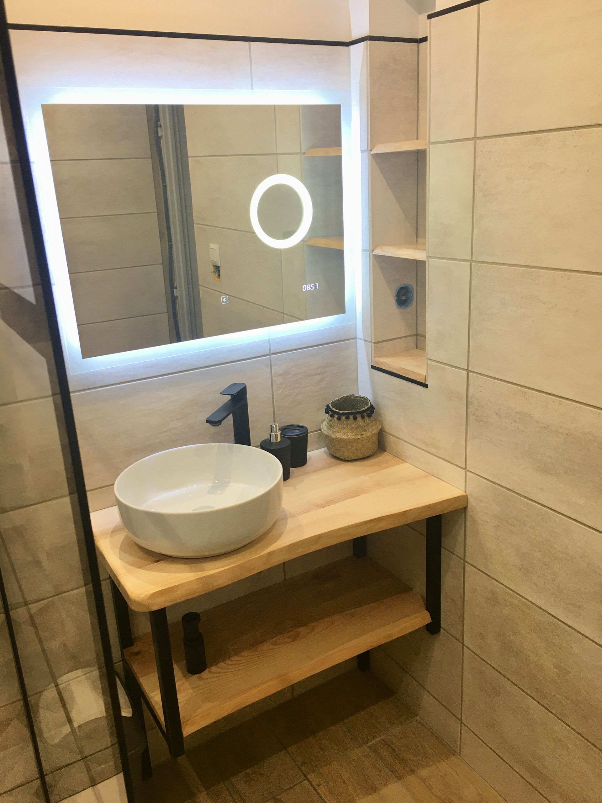 Meuble-lavabo moderne avec vasque ronde, robinet noir, miroir éclairé et étagère en bois.
