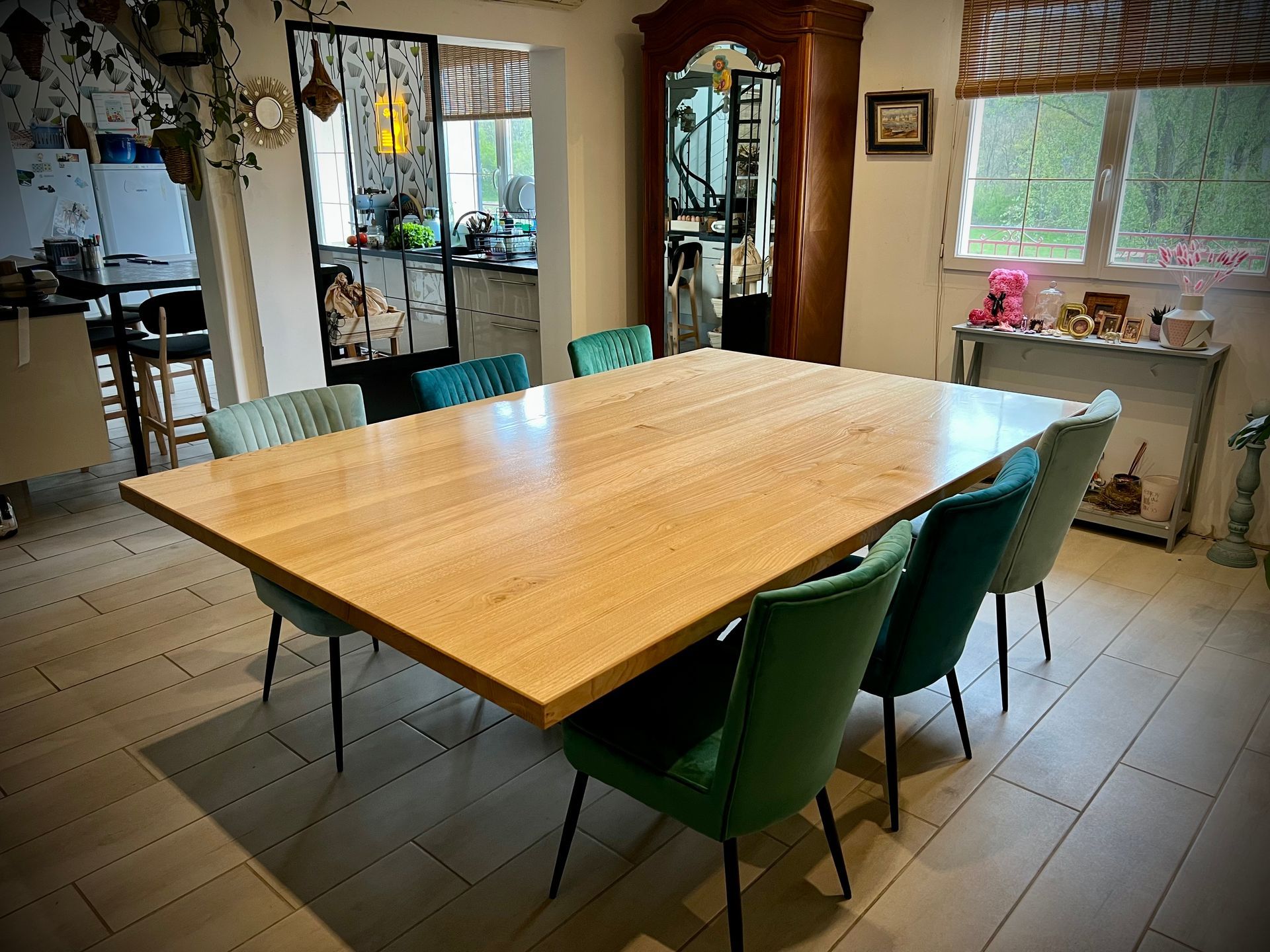 Salle à manger avec une grande table en bois, des chaises vertes et de la lumière naturelle entrant par les fenêtres.