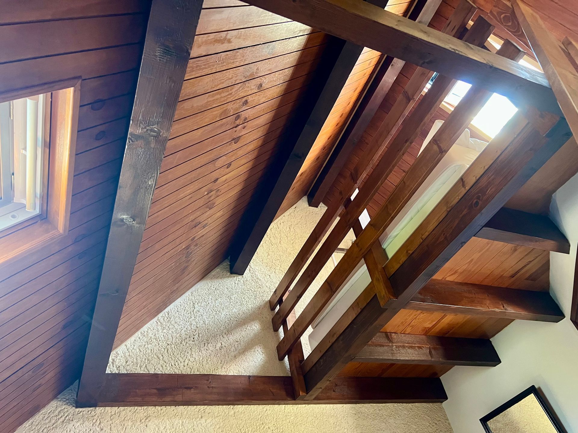 Escalier en bois vu d'en bas, avec moquette beige et rampes foncées sous un plafond en bois incliné