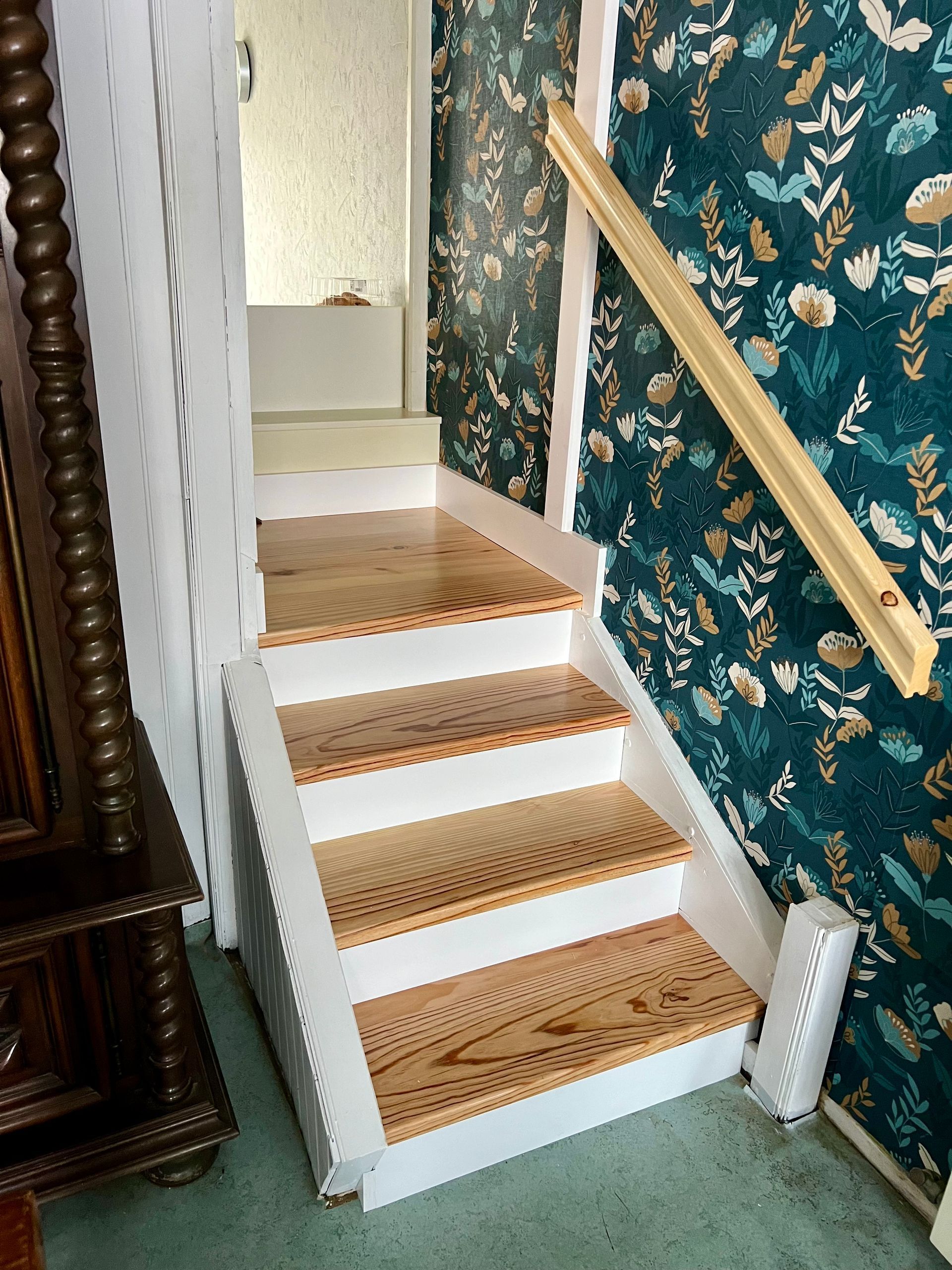 Escalier étroit recouvert de moquette, avec des moulures blanches et une rampe en laiton, à côté d'un papier peint floral turquoise.