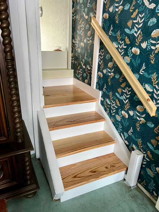 Escalier étroit avec marches beiges tressées, moulures blanches et rampe en laiton, à côté d'un papier peint à motifs floraux.