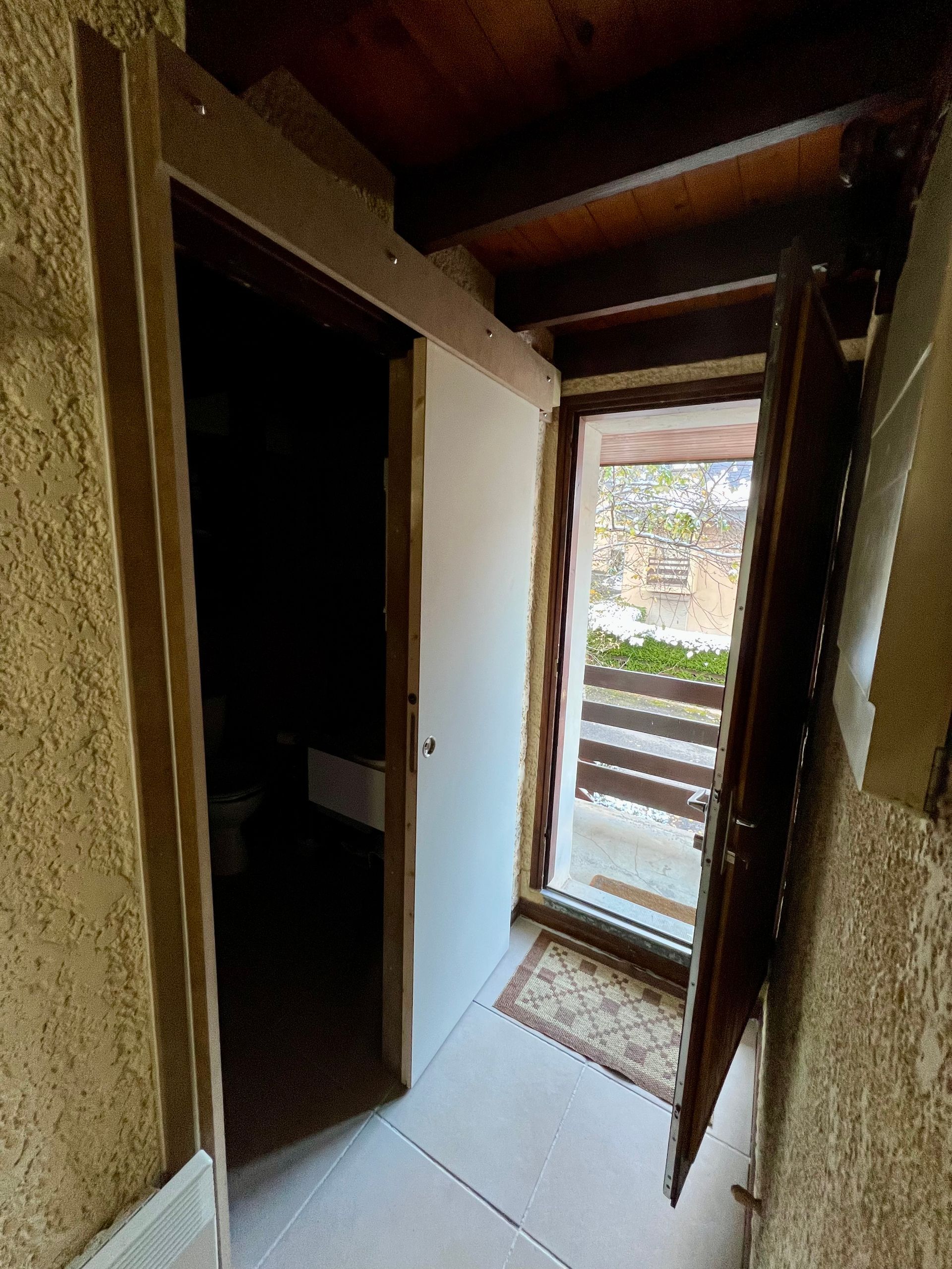 Couloir étroit avec porte ouverte donnant sur un balcon ou une terrasse extérieure lumineuse.