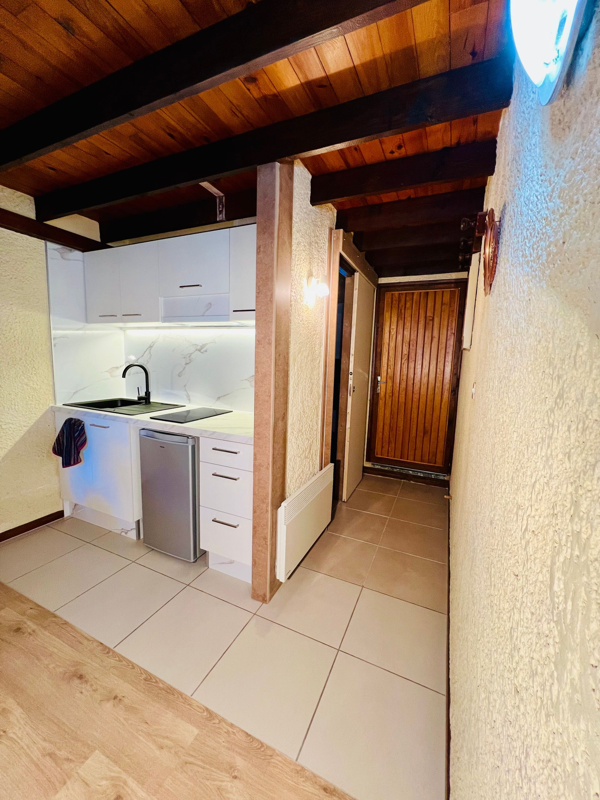 Petite cuisine avec placards blancs, sol carrelé, poutres apparentes et couloir menant à une porte en bois.