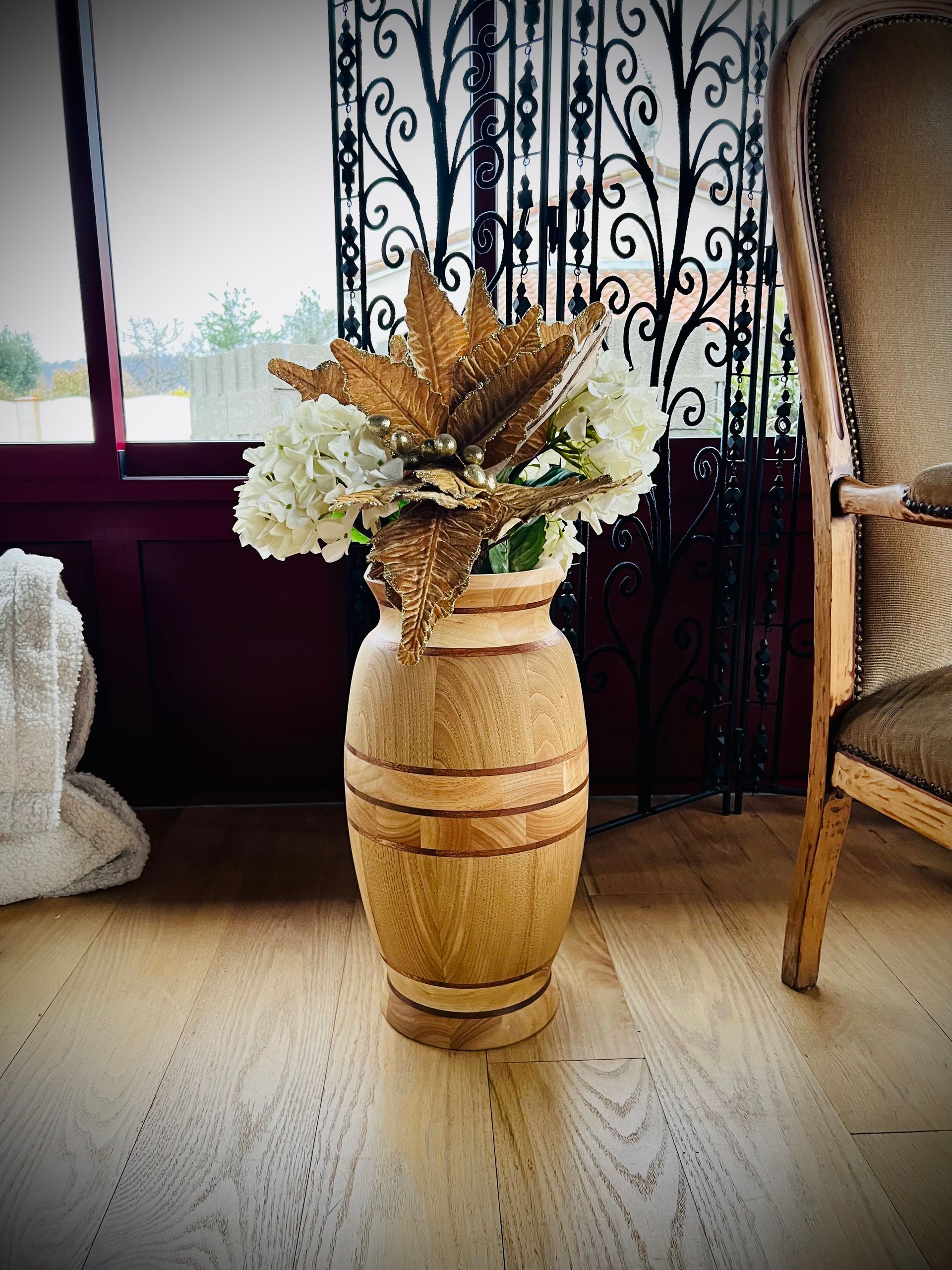 Grand vase tressé contenant des fleurs séchées dans une pièce lumineuse, à côté d'une chaise et d'une fenêtre.