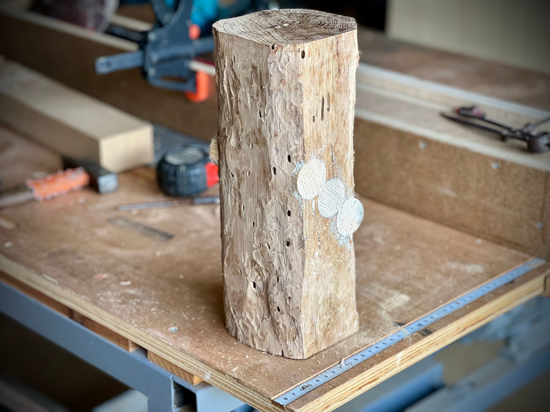 Une bûche de bois brut posée sur un établi dans un atelier, avec des outils et des morceaux de bois en arrière-plan.