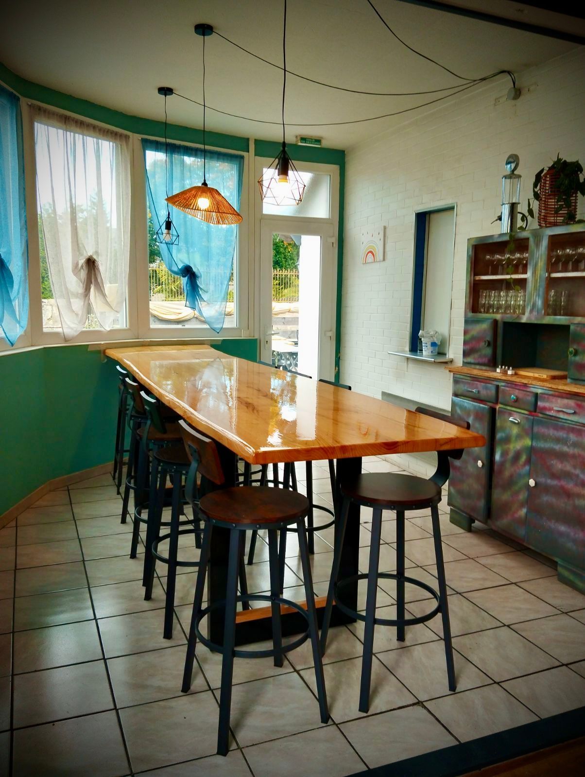 Petite cuisine avec une table en bois, des tabourets, un sol carrelé et des fenêtres laissant entrer la lumière du soleil.