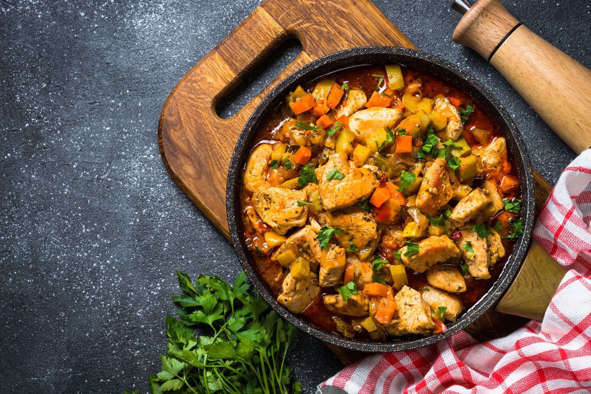 Una sartén con guisado de pollo con zanahorias y perejil sobre una tabla de cortar.