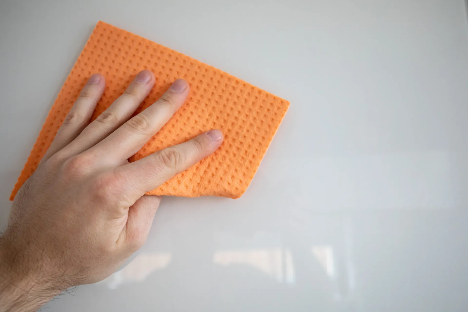 Mano limpiando una superficie blanca con un paño de limpieza naranja.