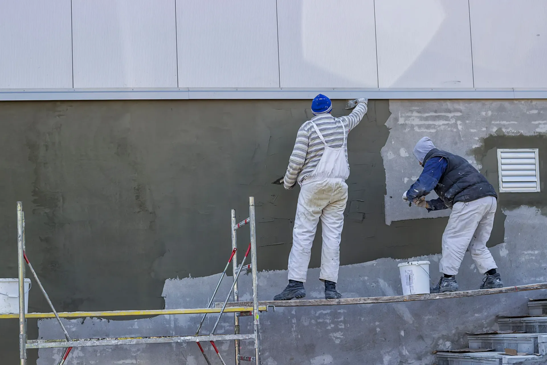 Dos trabajadores enyesan la pared exterior de un edificio con material gris, usando llanas.
