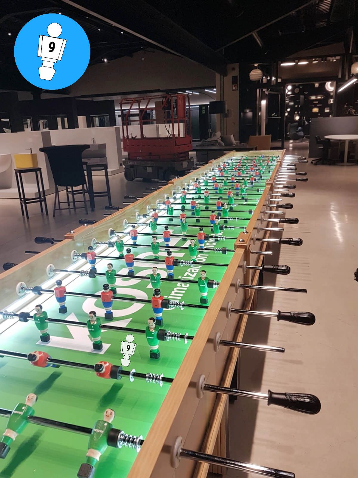 Una mesa de futbolín muy larga, verde y luminosa, en un espacio interior con ascensor.