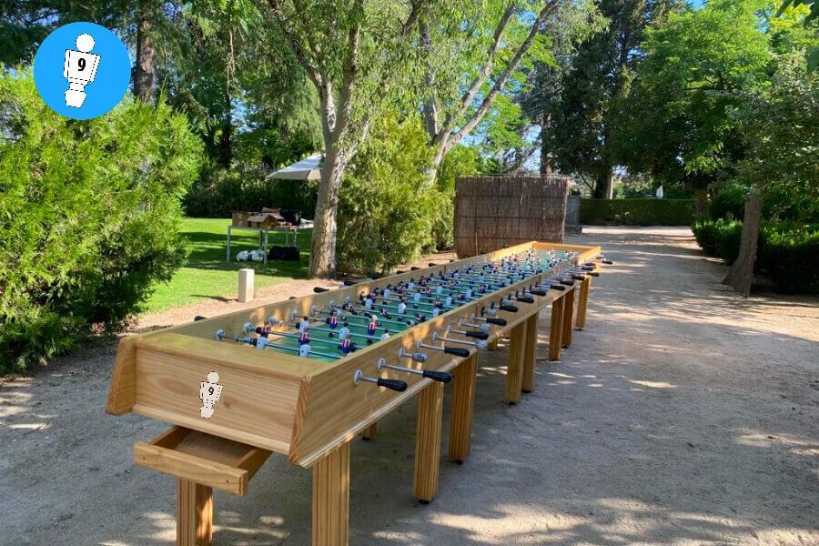 Mesa de futbolín larga sobre un camino de grava en un parque, con jugadores en las barras. Superficie de juego verde, estructura y patas de madera clara.