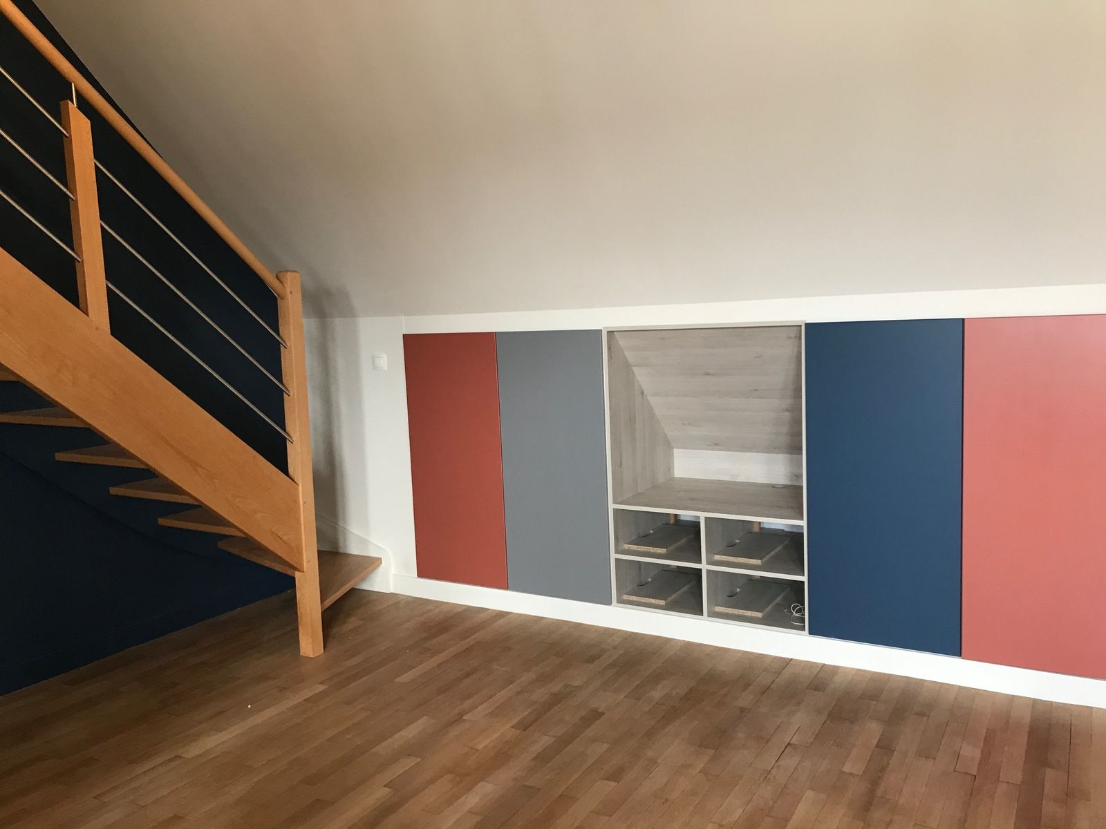 Un escalier en bois jouxte des placards intégrés aux portes rouges, grises et bleues, dans une pièce au parquet en bois dur.