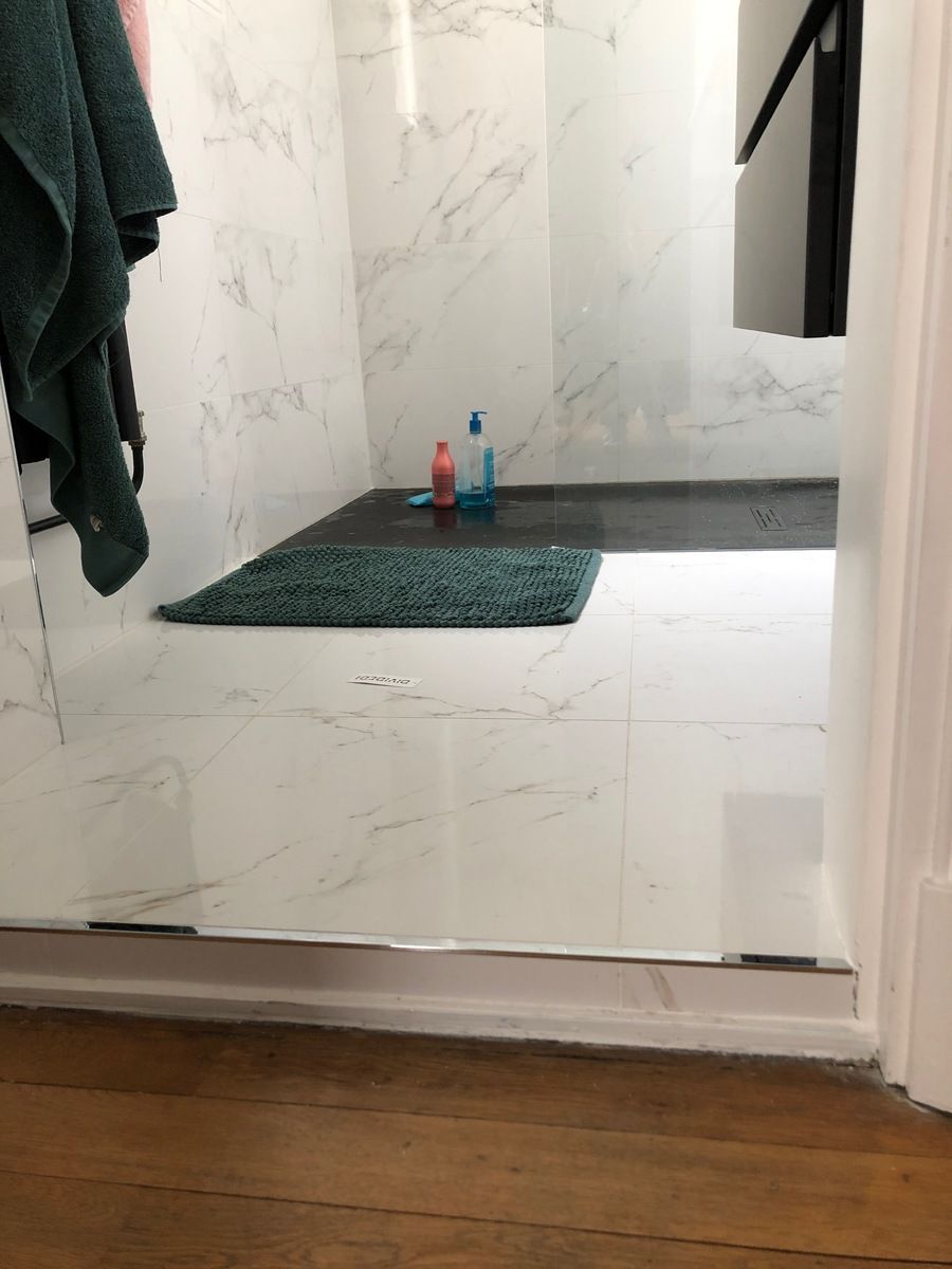 Intérieur de douche avec porte en verre, carrelage blanc et tapis de bain vert. Sol en bois foncé.