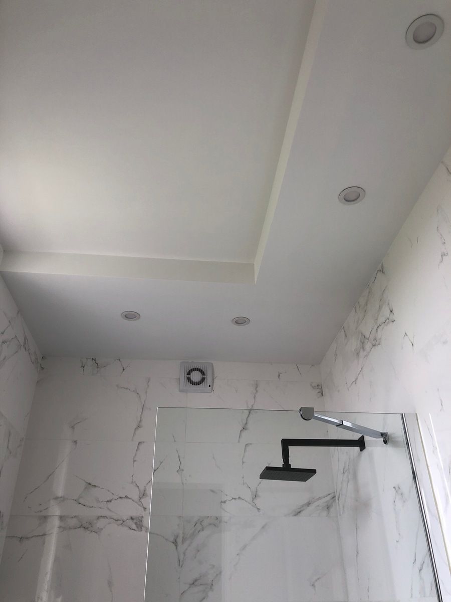 Plafond de salle de bains carrelé de blanc avec éclairage encastré et pommeau de douche derrière une paroi de verre.