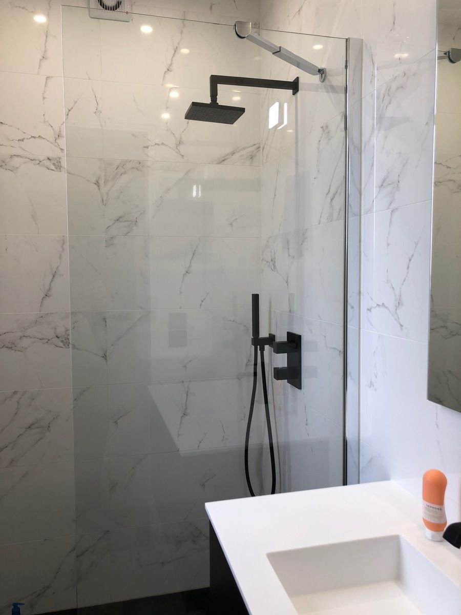 Douche avec parois à motif marbre, porte en verre, pommeau de douche noir, robinet et lavabo blanc.