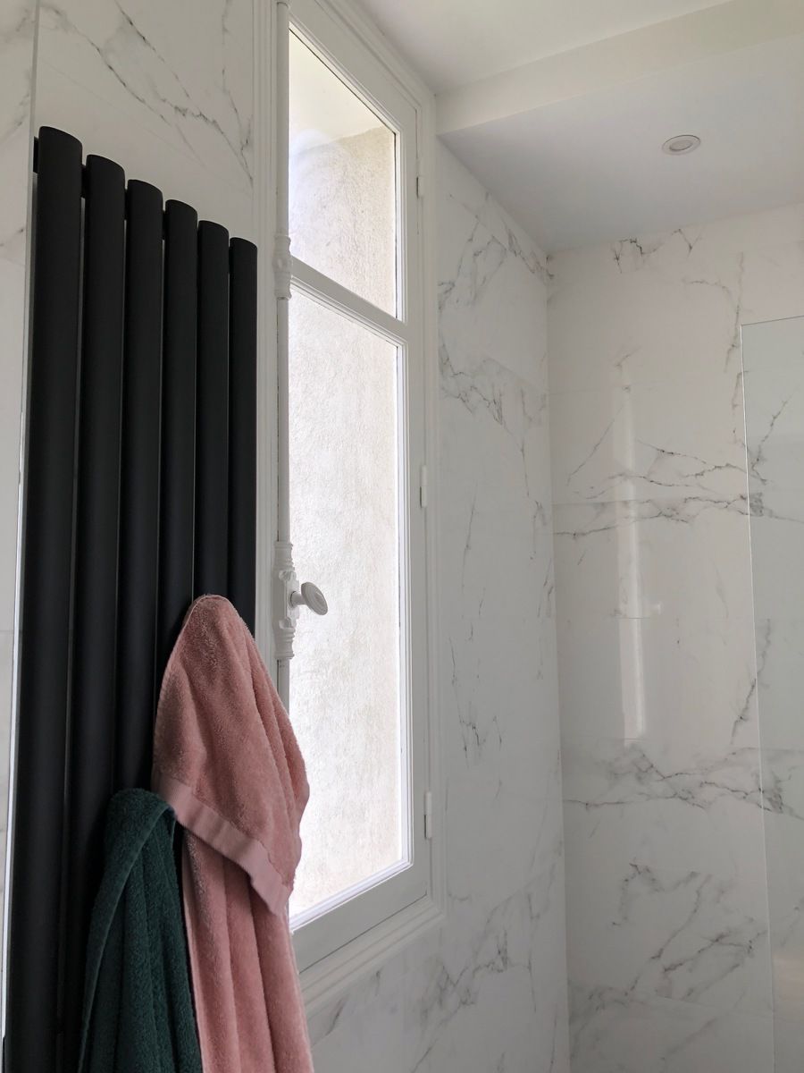 Radiateur noir avec des serviettes suspendues près d'une fenêtre dans une salle de bains carrelée de marbre.