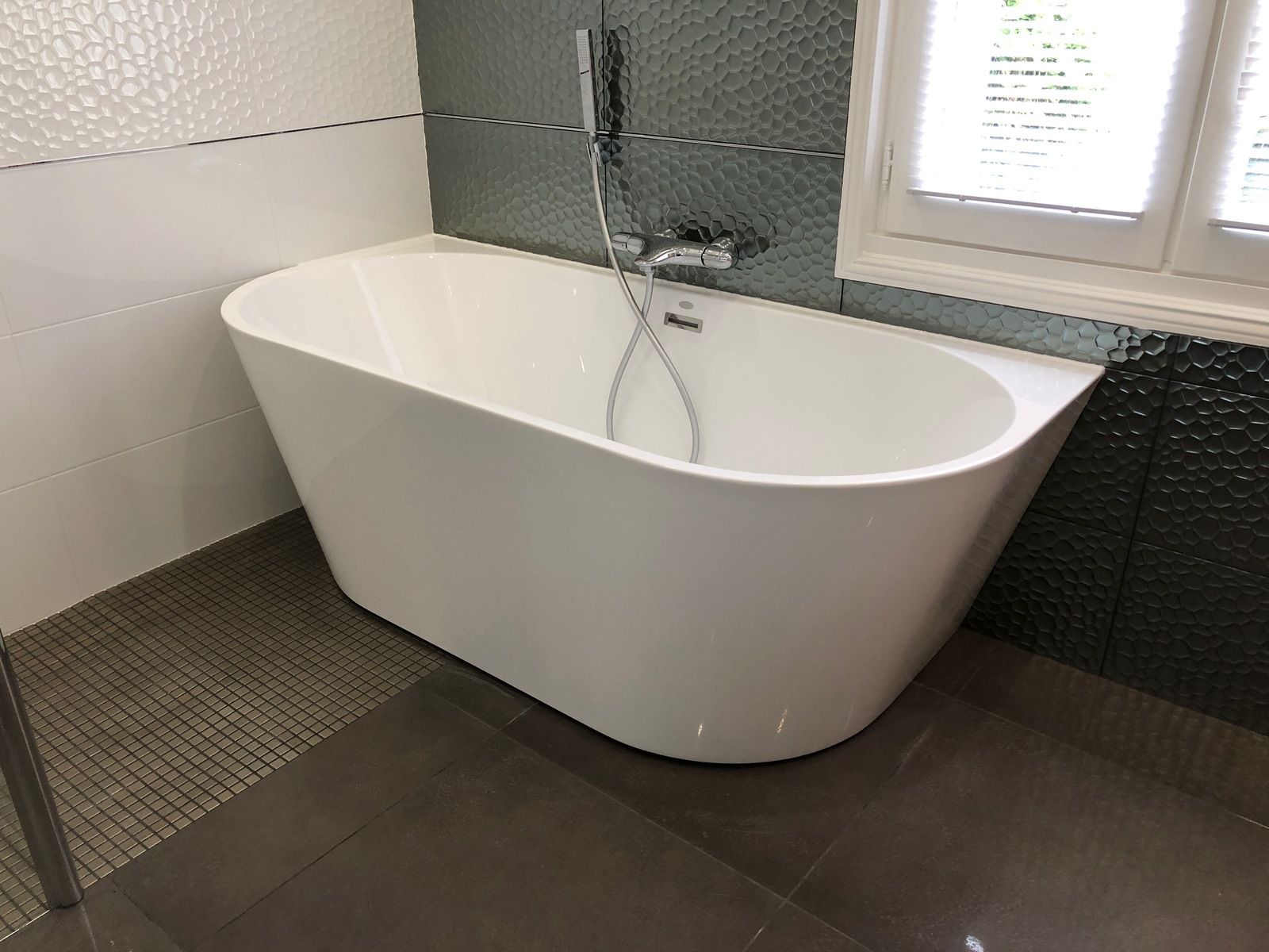 Baignoire blanche sur pieds dans une salle de bains, avec des carreaux foncés et une fenêtre.