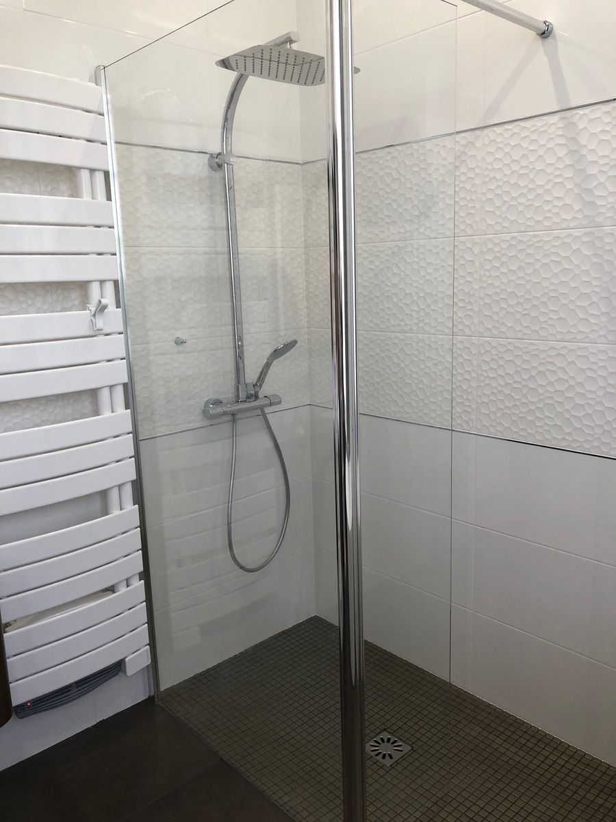 Cabine de douche avec paroi en verre, robinetterie chromée et carrelage blanc texturé.