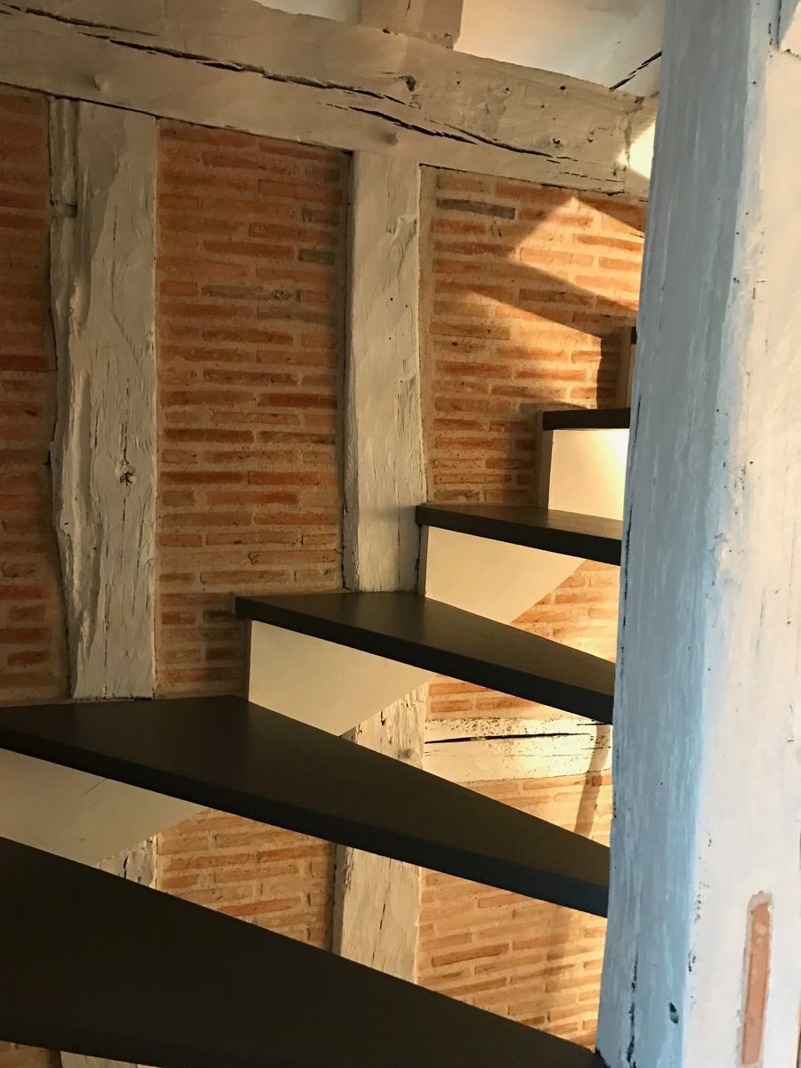 Escalier en bois avec marches noires, contremarches blanches et mur en briques rouges, avec des poutres de soutien en bois vieilli.