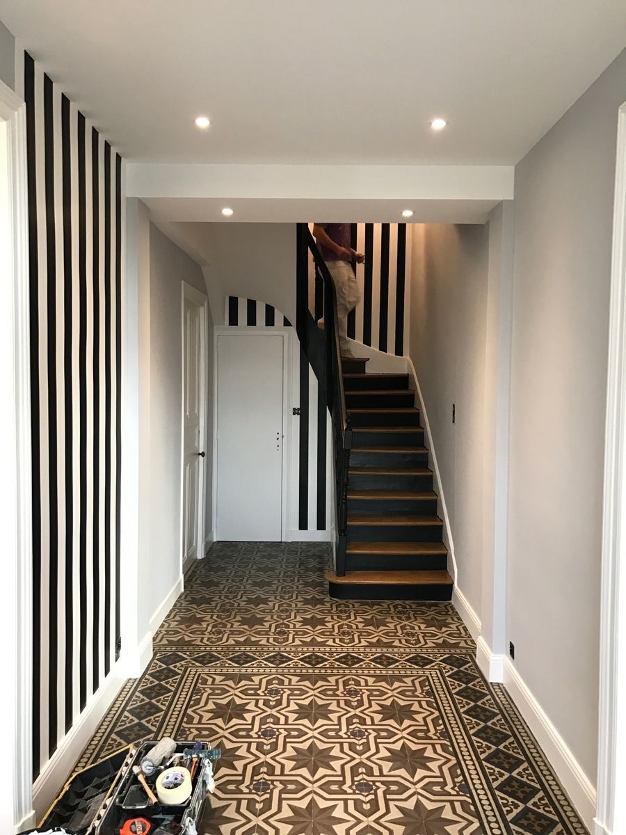 Couloir avec mur à rayures noires et blanches, sol à motifs et escalier. Une personne monte les escaliers.