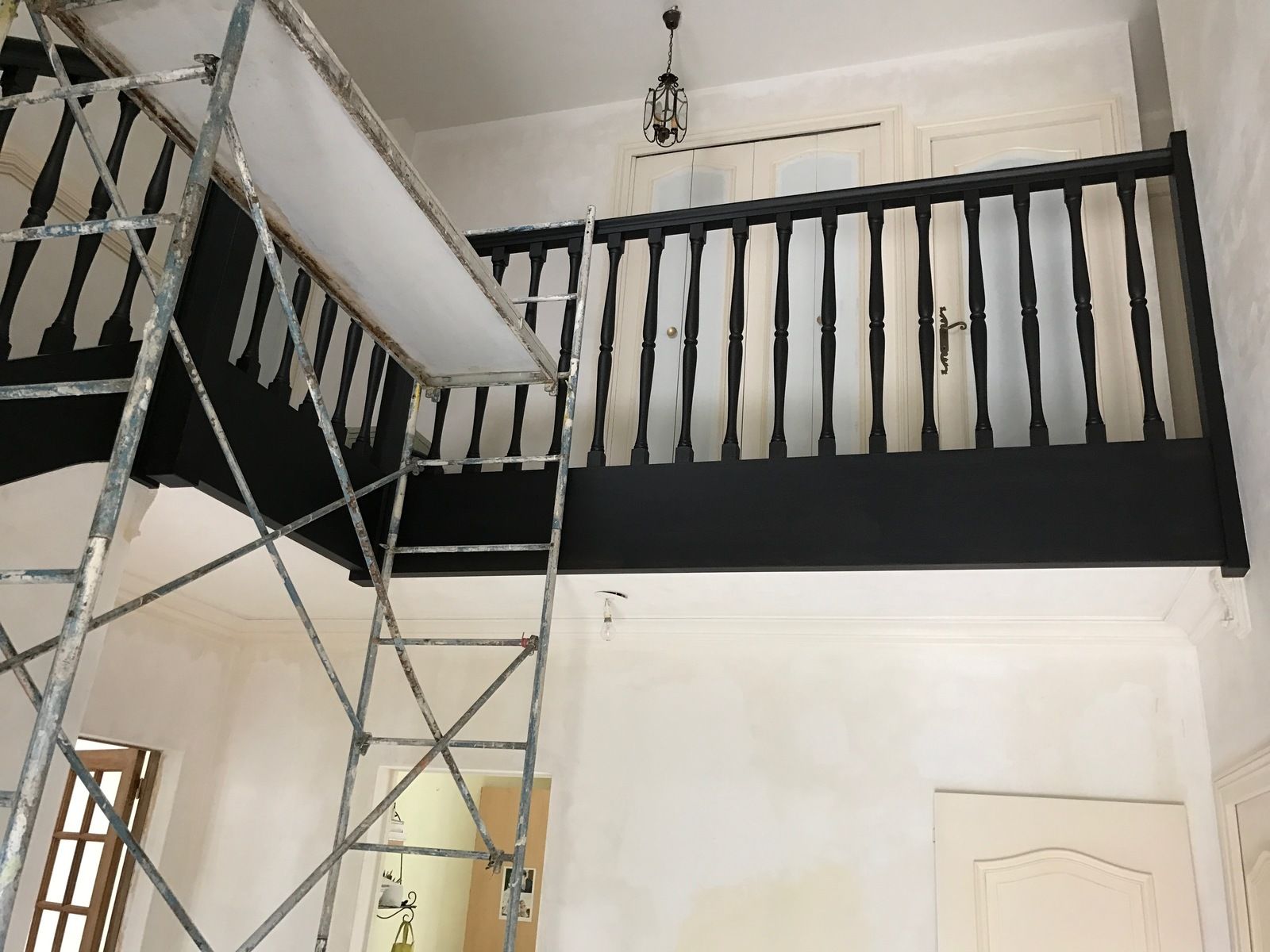 Rambarde noire sur le palier du deuxième étage, avec échafaudages en dessous. Murs et porte blancs.