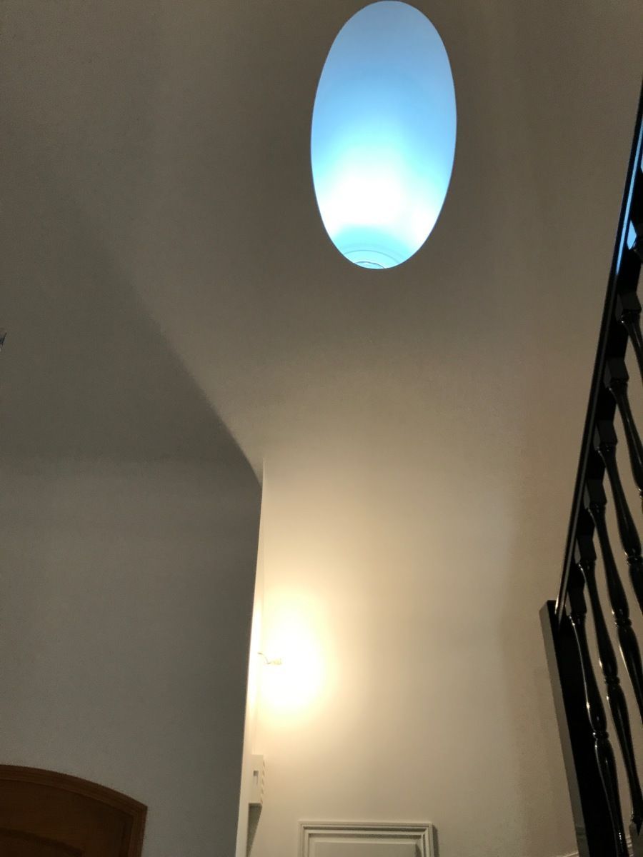 Un luminaire ovale fixé au plafond éclaire un mur blanc. Rambarde noire à droite.