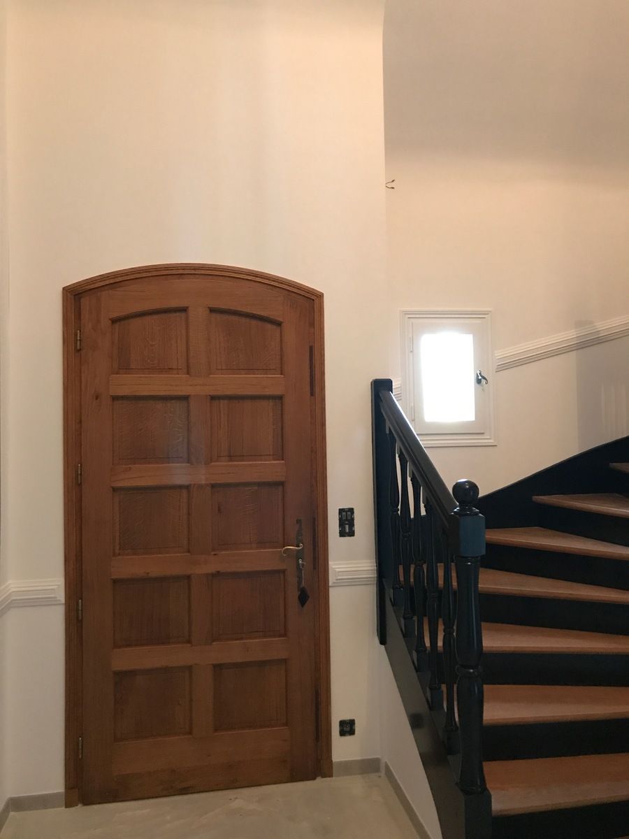 Porte cintrée en bois à côté d'un escalier. L'escalier a une rampe noire, des marches brunes et une petite fenêtre.