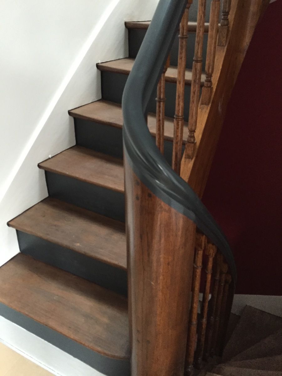 Un escalier en bois aux contremarches peintes en gris foncé et aux marches brunes est visible. On aperçoit une rampe en bois courbée avec des balustres.