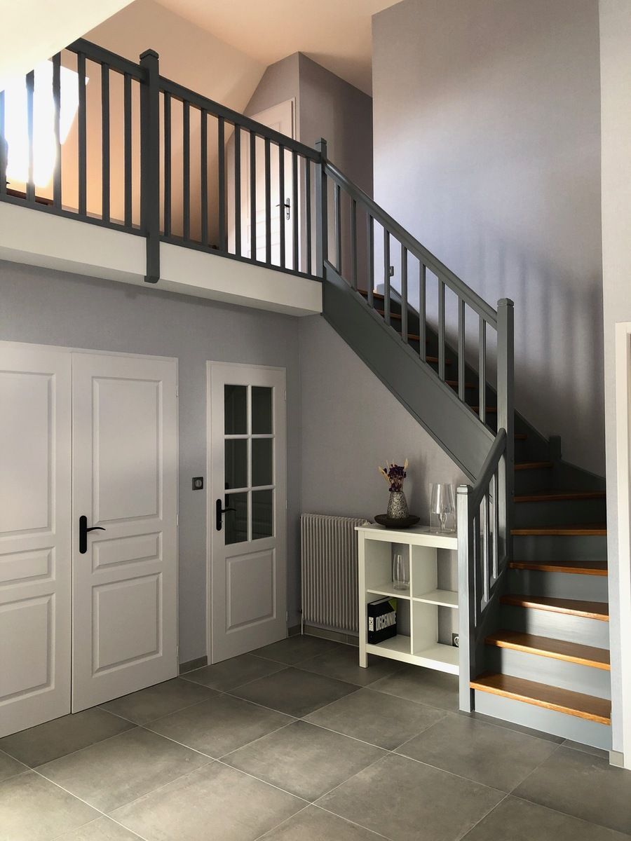 Vue intérieure d'une maison avec un escalier, un palier, deux portes et une étagère blanche. Dominante de gris et de blanc.
