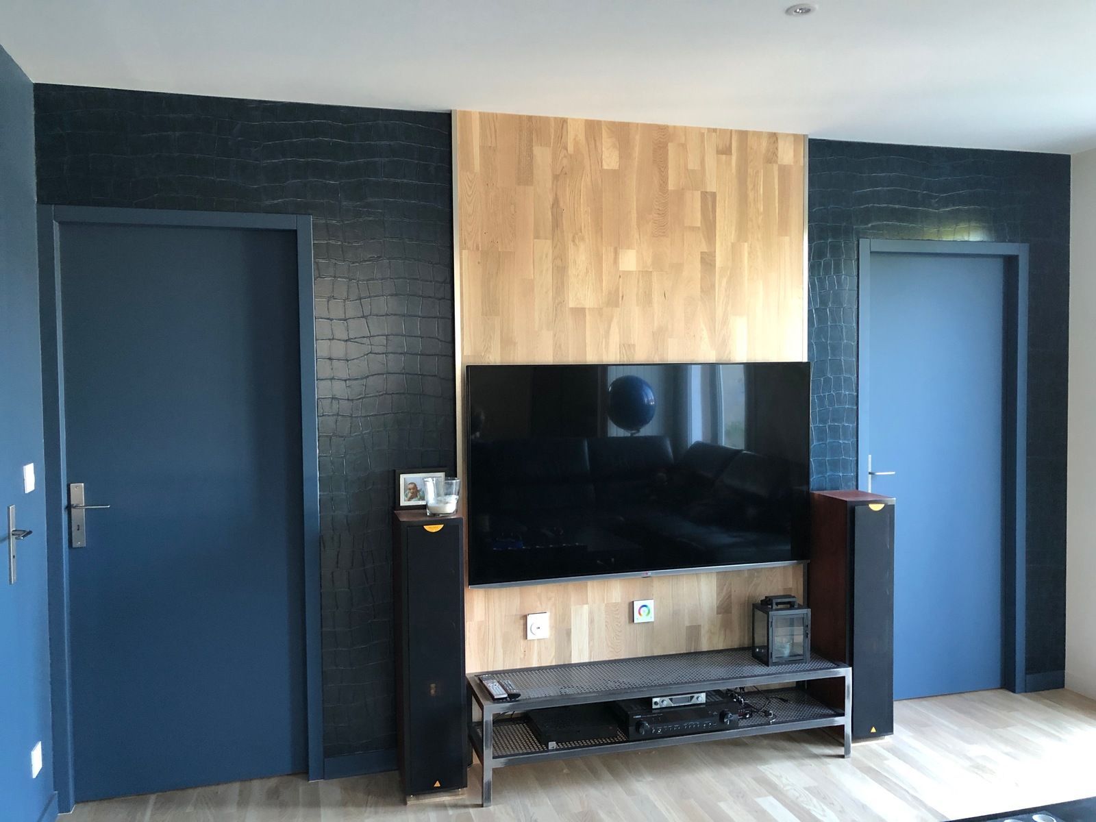 Salon avec télévision sur panneau de bois, flanqué de deux portes bleues et de murs à la texture sombre.