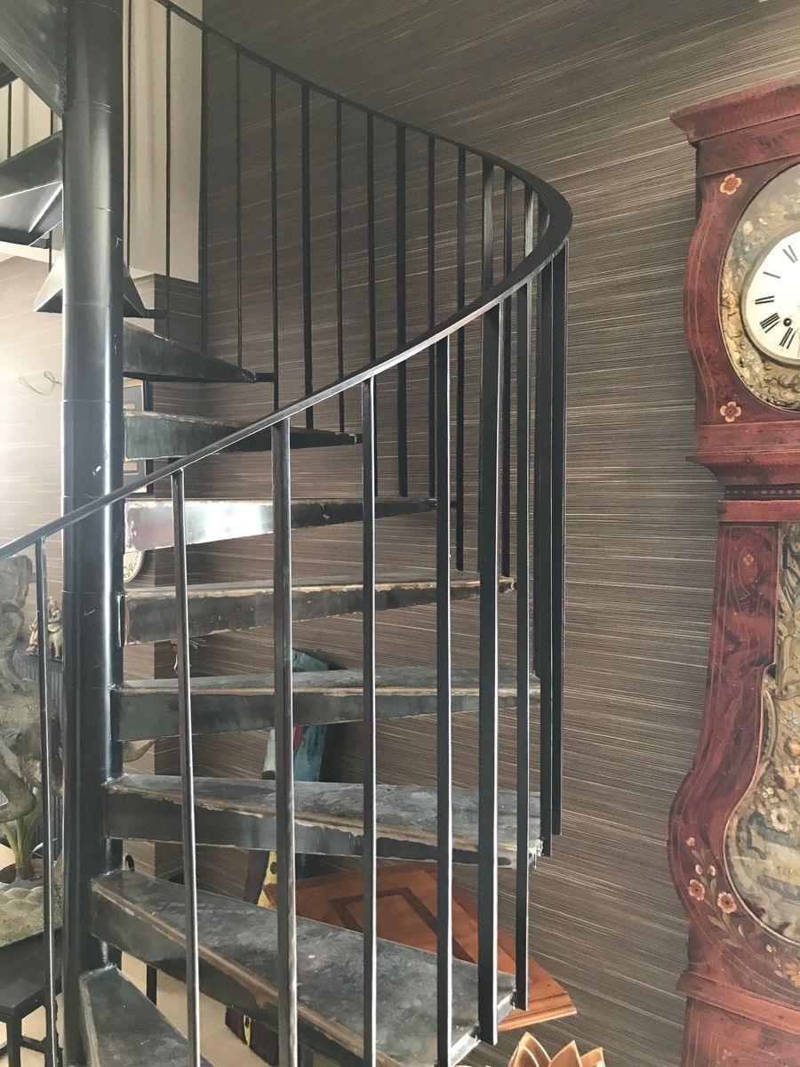 Escalier en colimaçon noir avec marches en bois foncé et rampes en métal noir, adossé à un mur texturé.