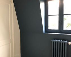 Vue intérieure avec un mur bleu-gris, une porte blanche, une fenêtre à cadre foncé et un radiateur foncé.