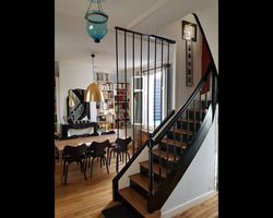 Escalier à rampe noire jouxtant un coin repas avec table, chaises et livres. Luminaires suspendus bleus et dorés.