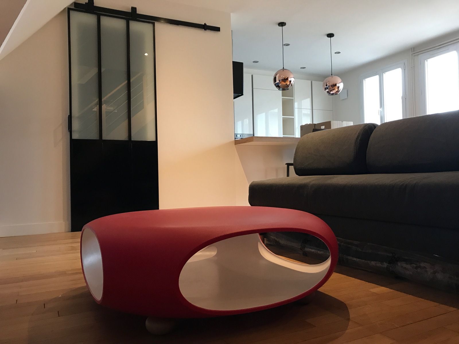 Table basse ovale rouge dans un salon moderne avec canapé noir et porte-fenêtre coulissante.