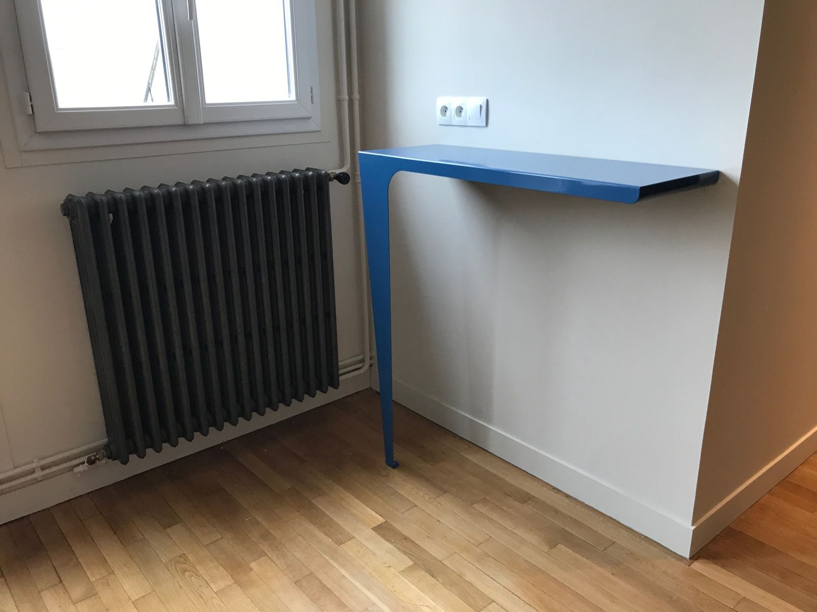 Bureau mural bleu dans une pièce avec radiateur et fenêtre. Sol en bois et murs blancs.