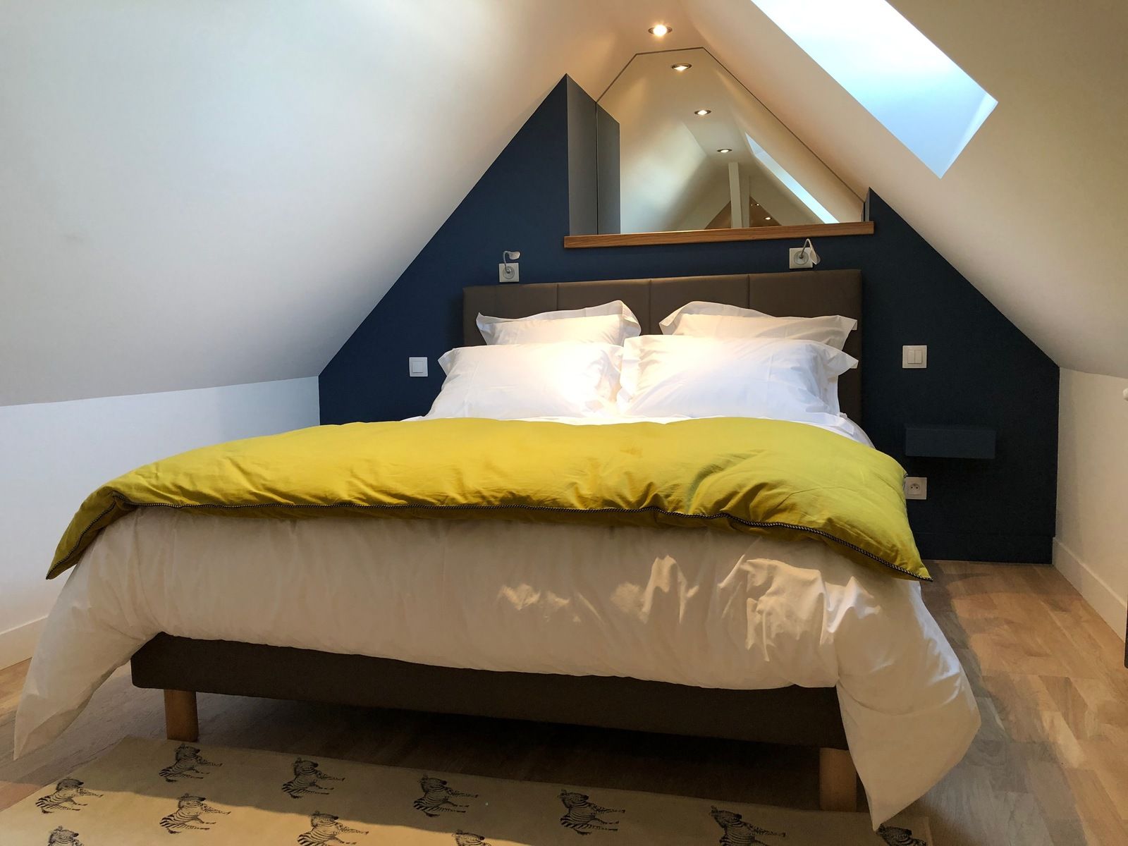 Chambre mansardée avec un lit, une couette jaune et un mur bleu derrière la tête de lit. Puits de lumière à droite.