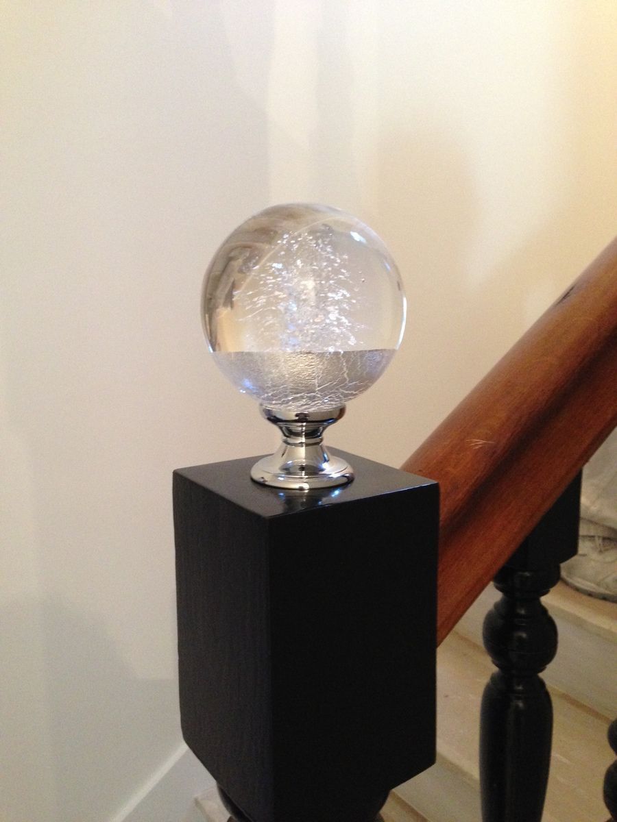 Poteau d'escalier noir surmonté d'un fleuron en verre transparent et d'une rampe en bois.