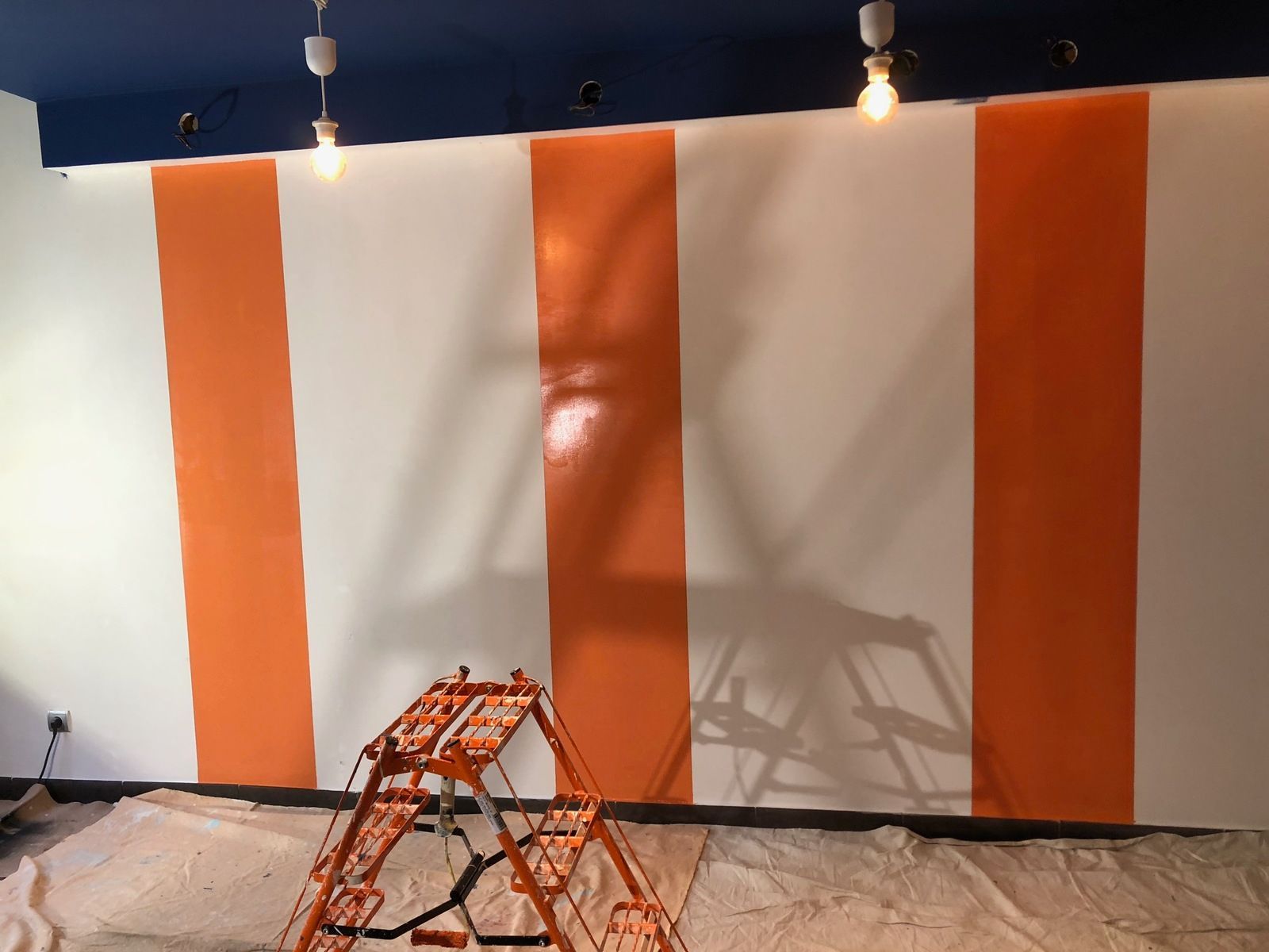 Mur à rayures orange et blanches avec du matériel de peinture au sol, sous un plafond bleu avec des suspensions lumineuses.