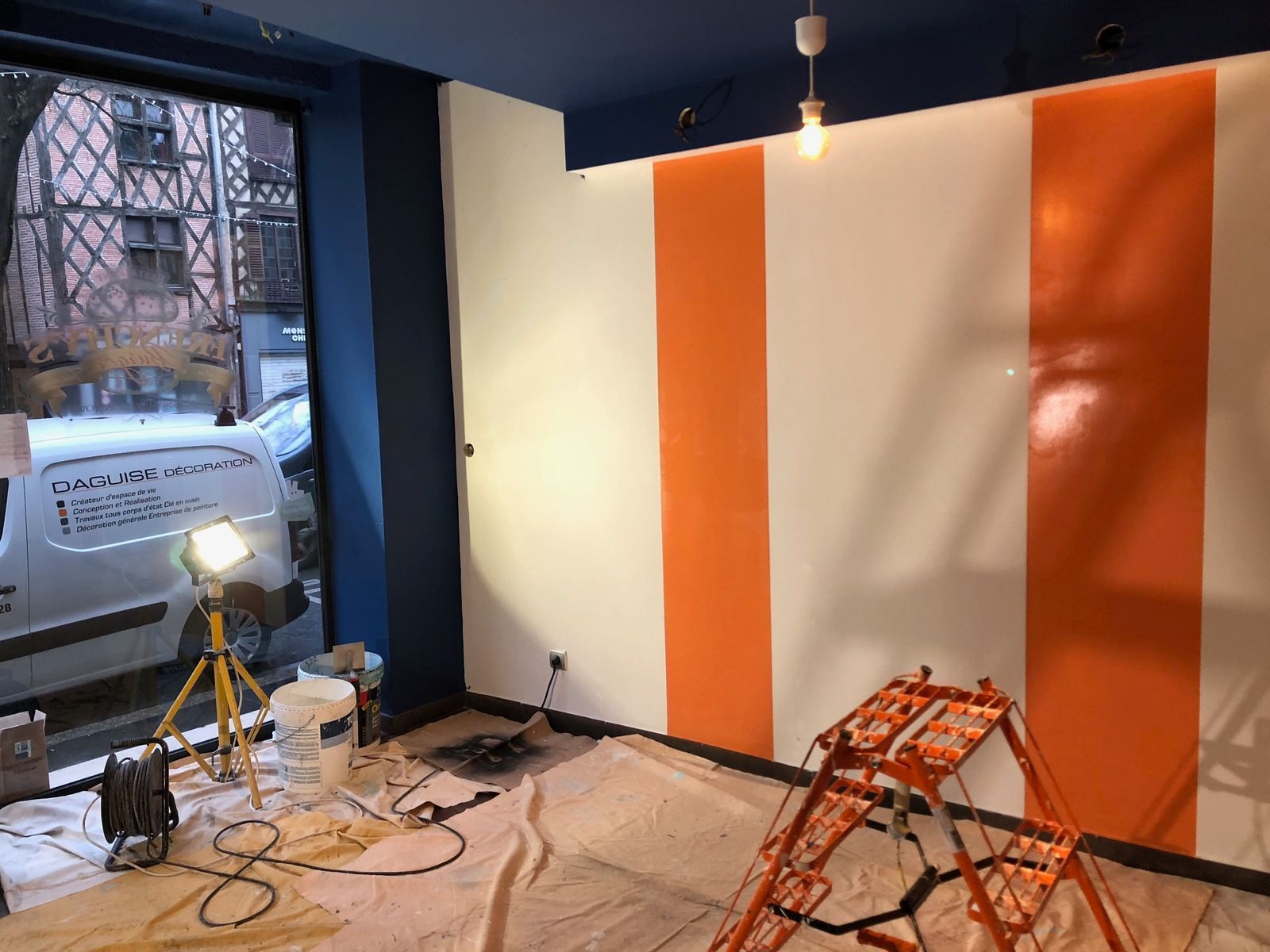 Mur intérieur peint à rayures orange et blanches ; échelle et matériel au sol.