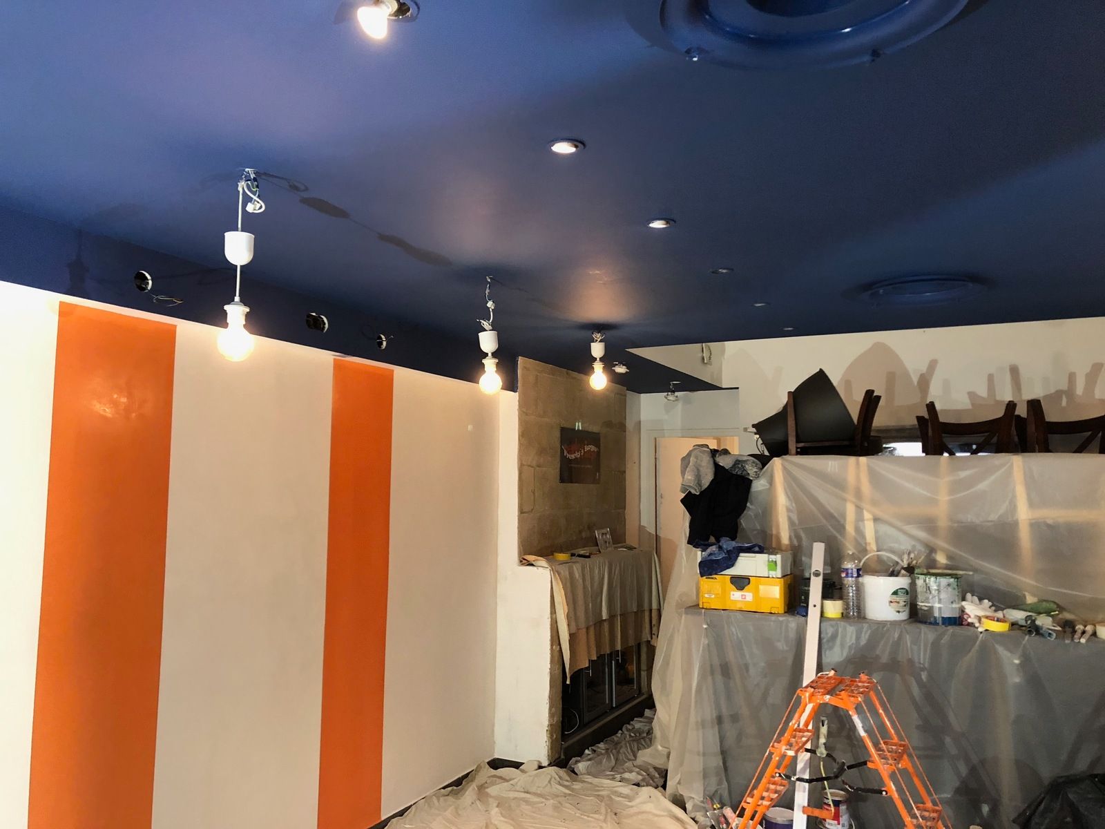 Intérieur aux murs rayés orange et blanc, plafond bleu et luminaires apparents. Travaux en cours.