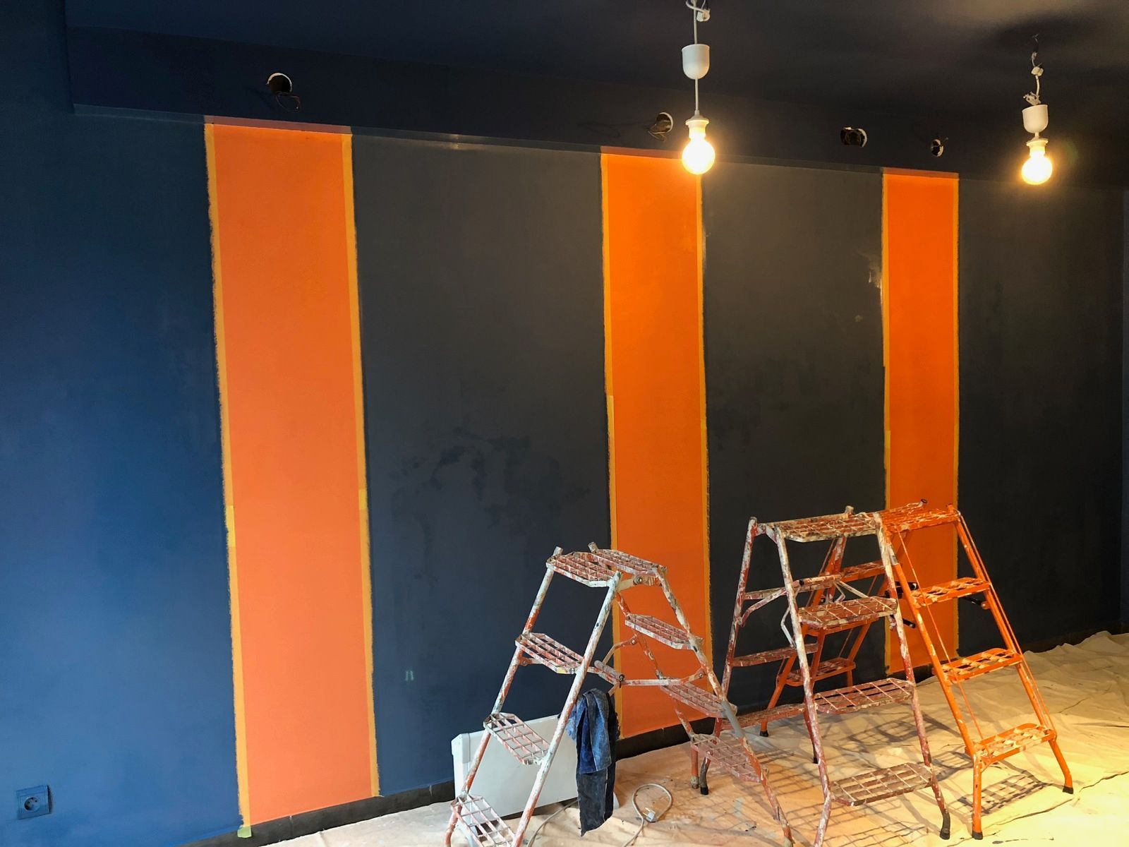 Mur intérieur en cours de peinture avec des rayures orange, deux échelles et des luminaires suspendus.