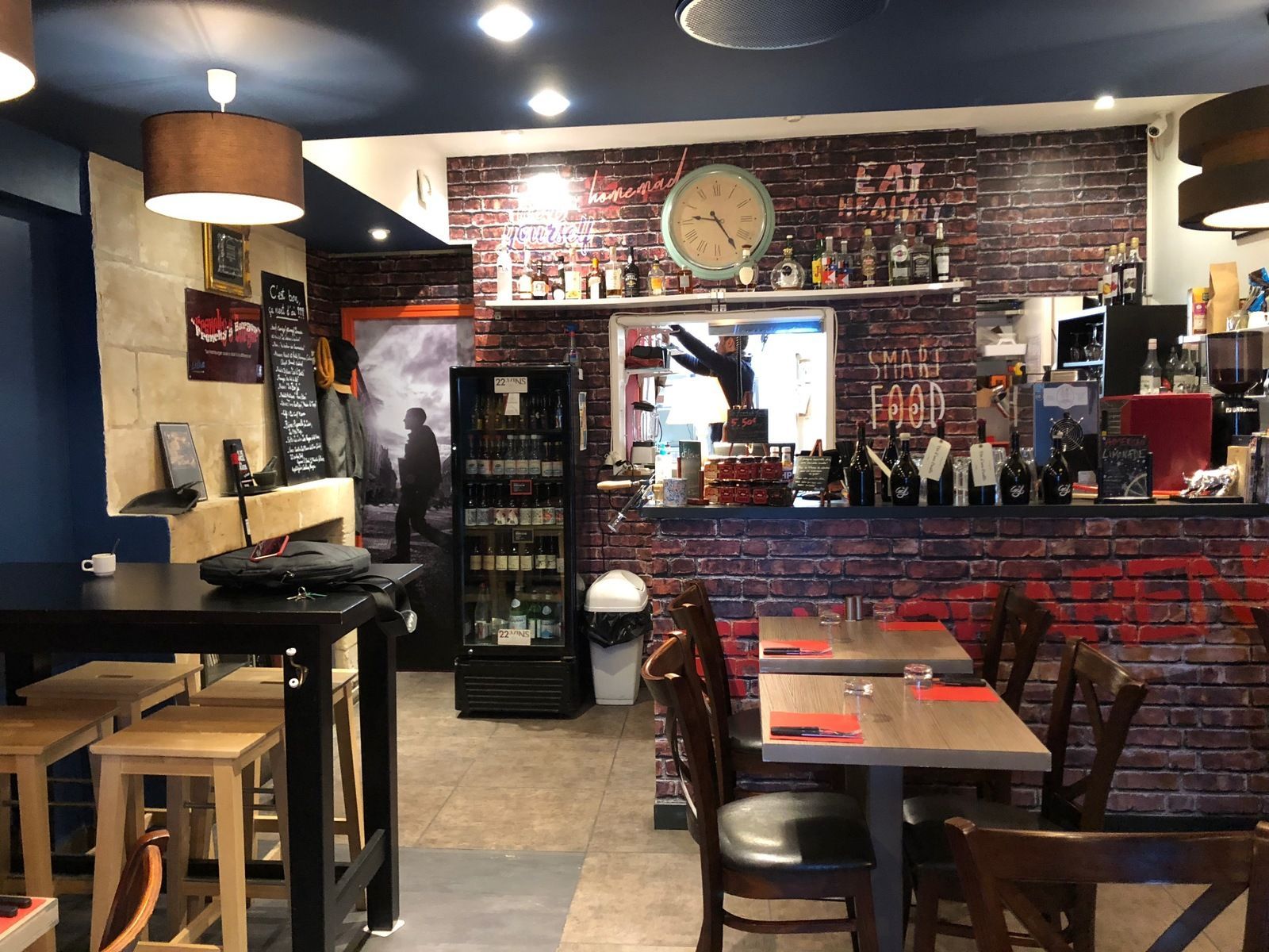 Vue intérieure d'un bistro avec tables, chaises, bar, mur de briques, horloge et réfrigérateur contenant des boissons.