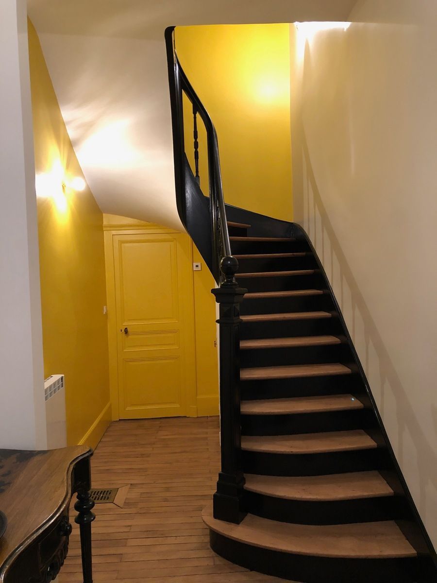 Escalier avec rampe noire, marches en bois et murs jaunes. Une porte jaune se trouve sur le palier.