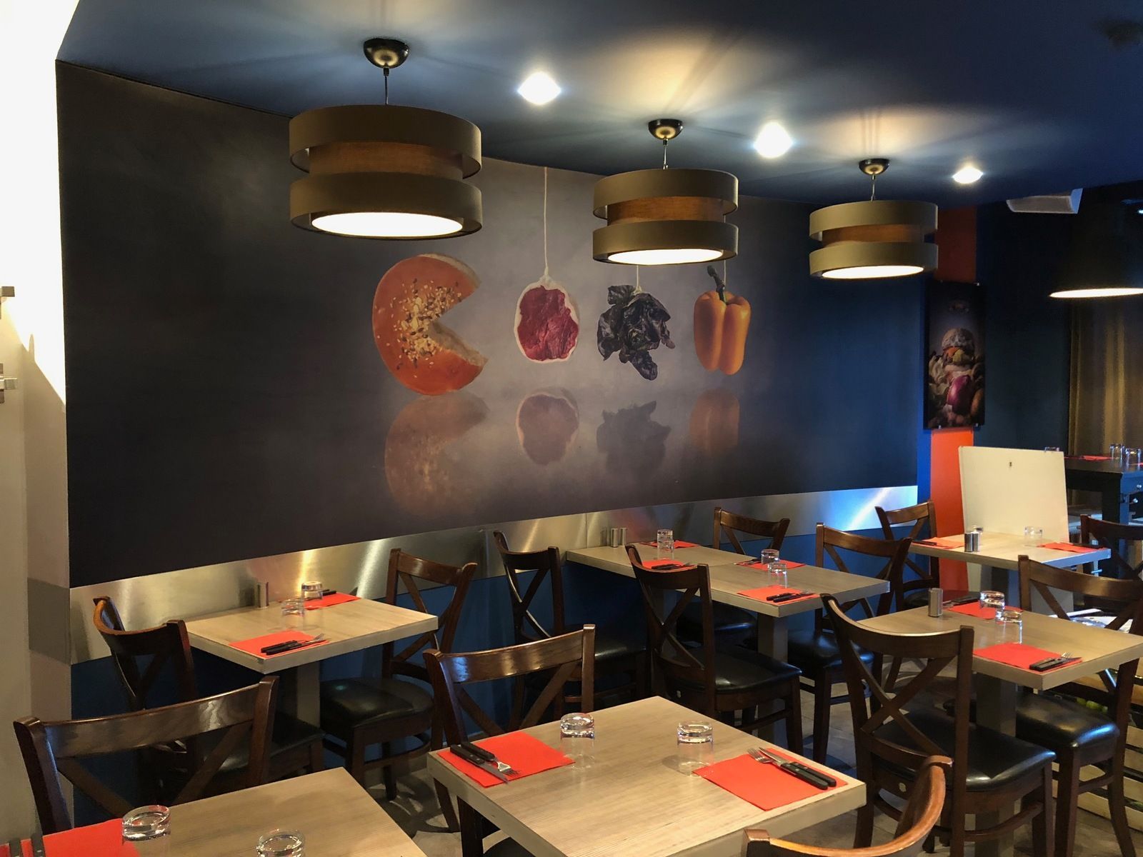 Intérieur d'un restaurant avec tables et chaises, une fresque murale représentant de la nourriture et des suspensions lumineuses.