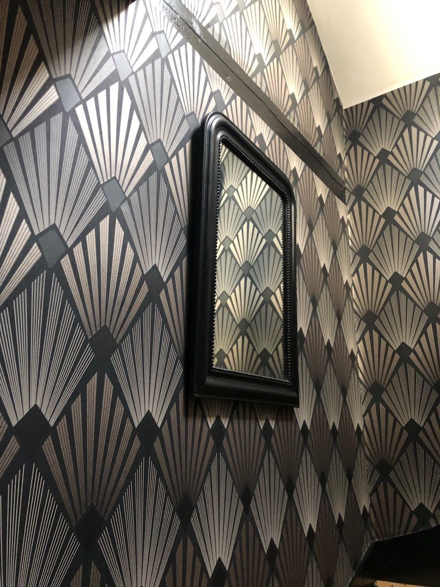 Miroir à cadre noir sur un mur recouvert d'un papier peint à motifs géométriques en forme d'éventail, dans les tons argent, gris et noir.