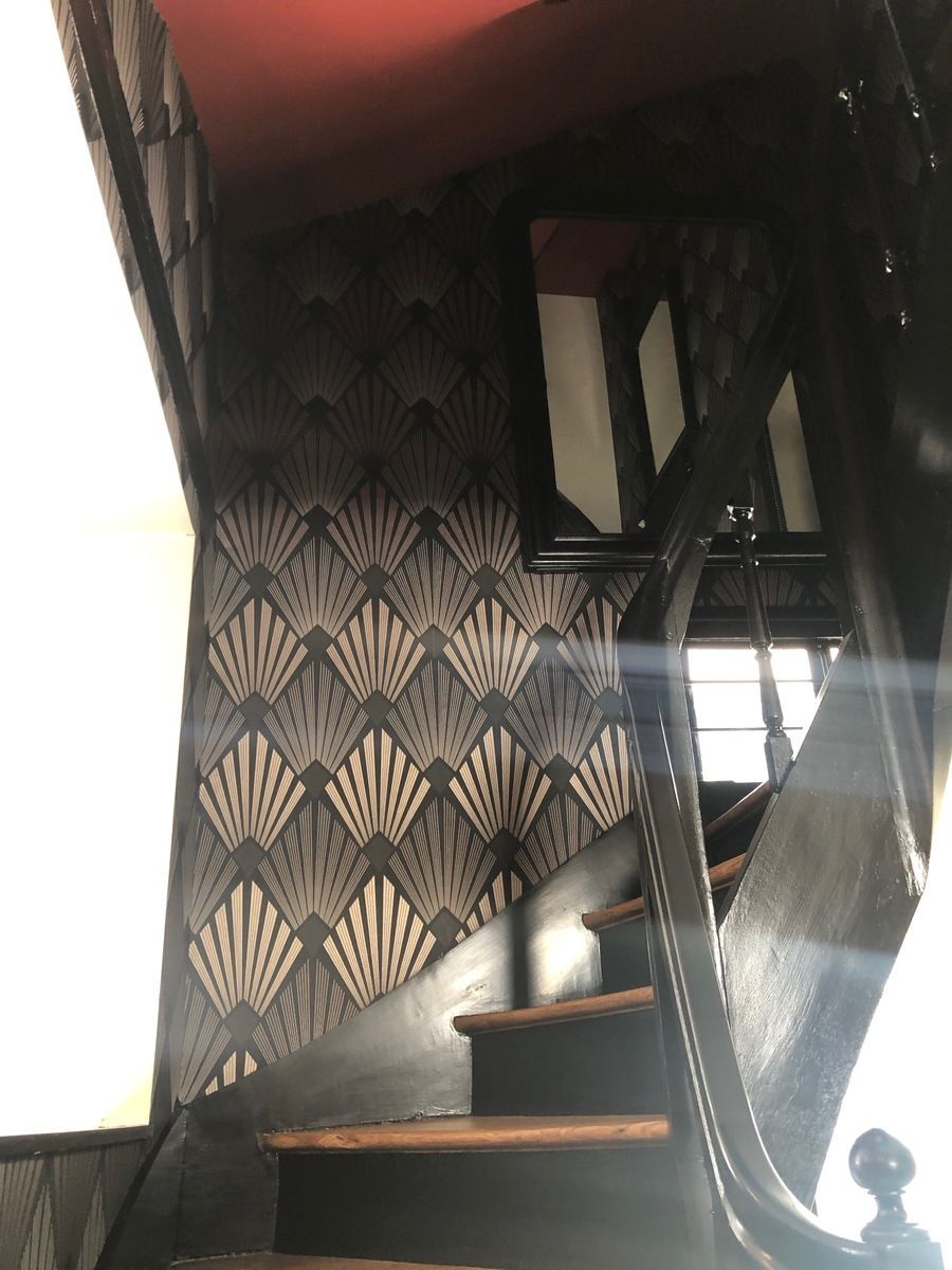 Escalier avec papier peint à motifs Art déco et rampe en bois foncé. Plafond rouge, lumière du soleil inondant la pièce.