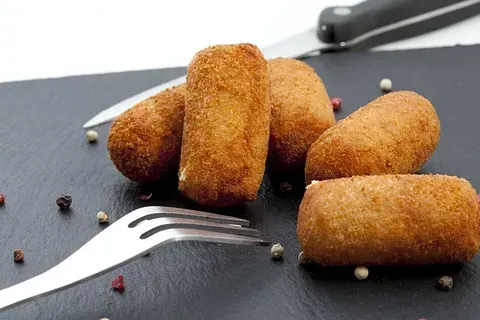 Croquetas doradas fritas sobre una superficie de pizarra negra con un tenedor, un cuchillo y granos de pimienta esparcidos.